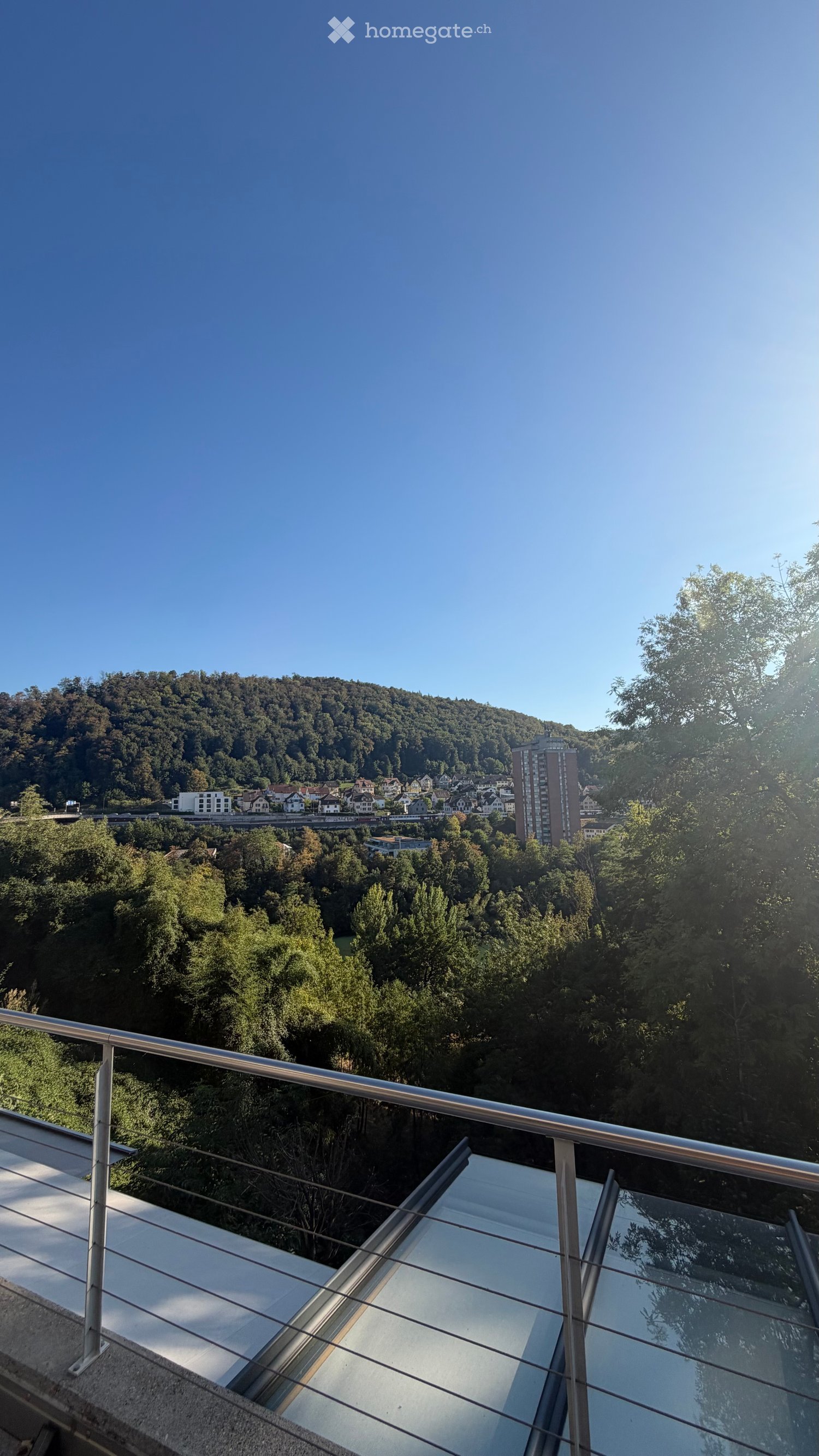 Balcony with glass railing, overlooking a forested area and town with buildings and a hill in the background.