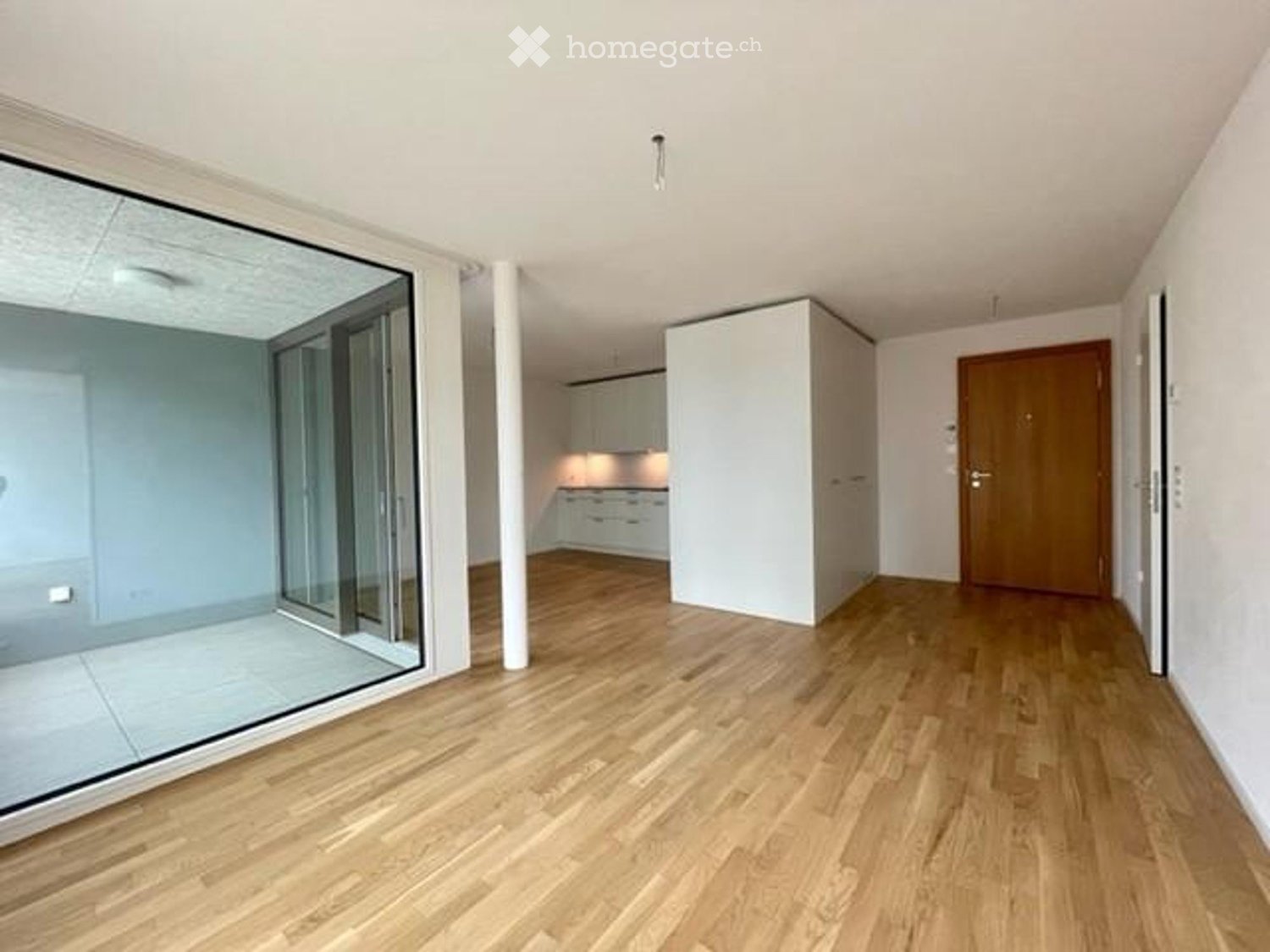 Empty hallway with wooden flooring, white walls, and a large glass sliding door leading to a balcony area. A closed wooden door is visible on the right side.