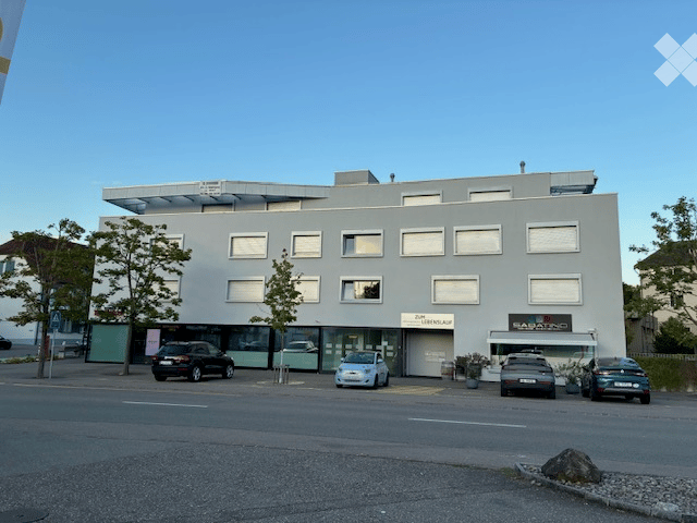 Modern multi-story building with a flat roof and large windows, featuring a storefront with signage and several parked cars in front.