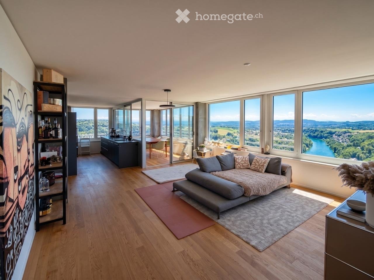 Open-concept living and dining area with large windows offering scenic views, a grey sectional sofa, a coffee table, a bookshelf, a kitchen island, and a dining table with chairs.