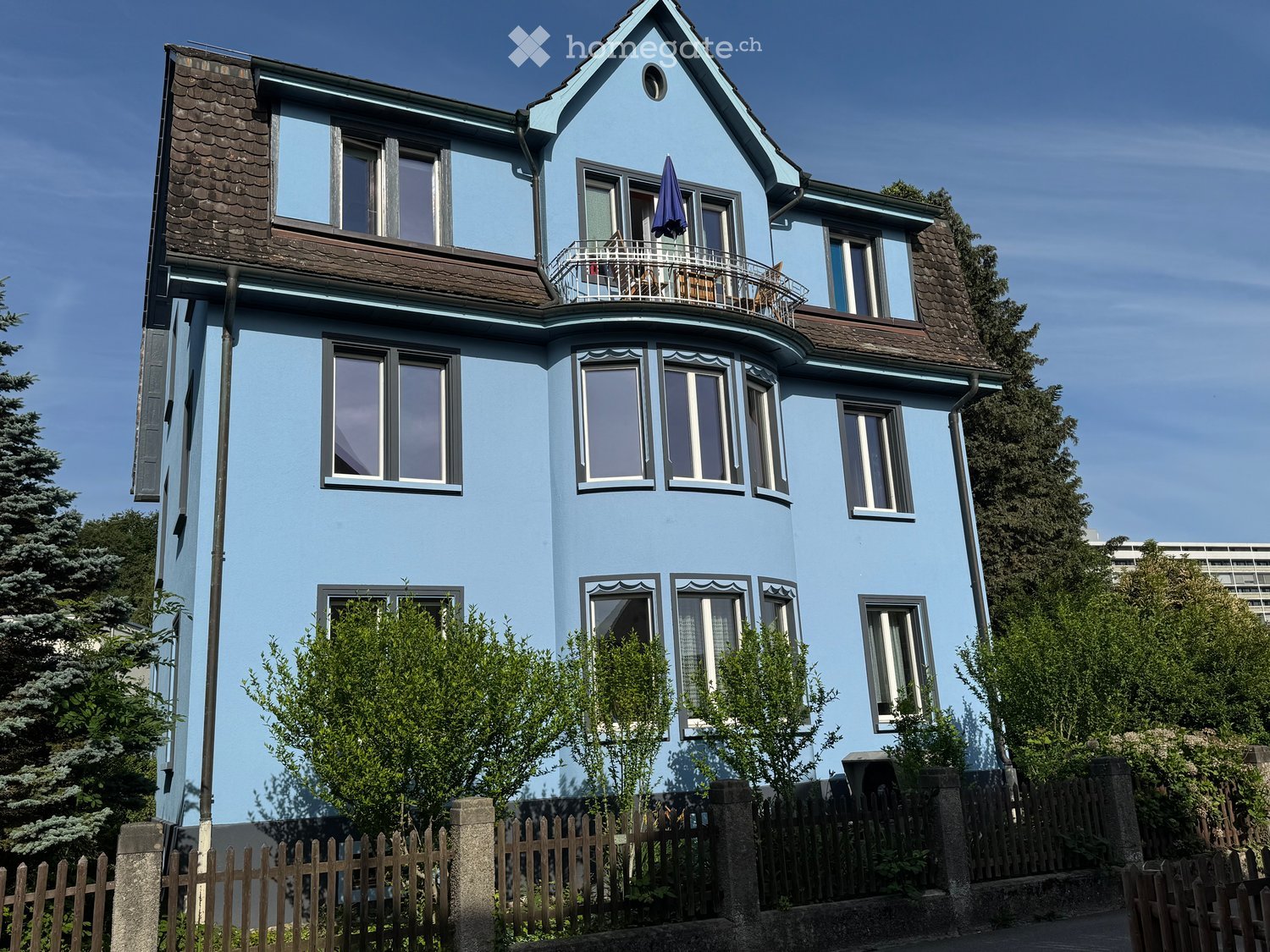Two-story blue house with multiple windows, a balcony on the upper floor, and a closed umbrella on the balcony railing. The house is surrounded by greenery and a wooden fence.