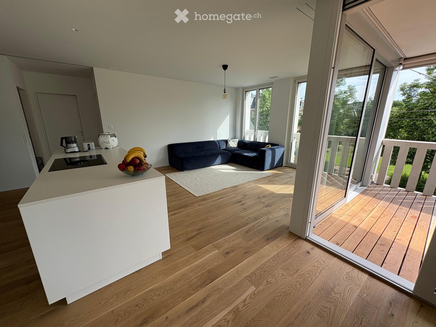 Modern living room with a kitchen island, dark blue sectional sofa, and large windows opening to a balcony. The room features light wooden flooring and a white rug.