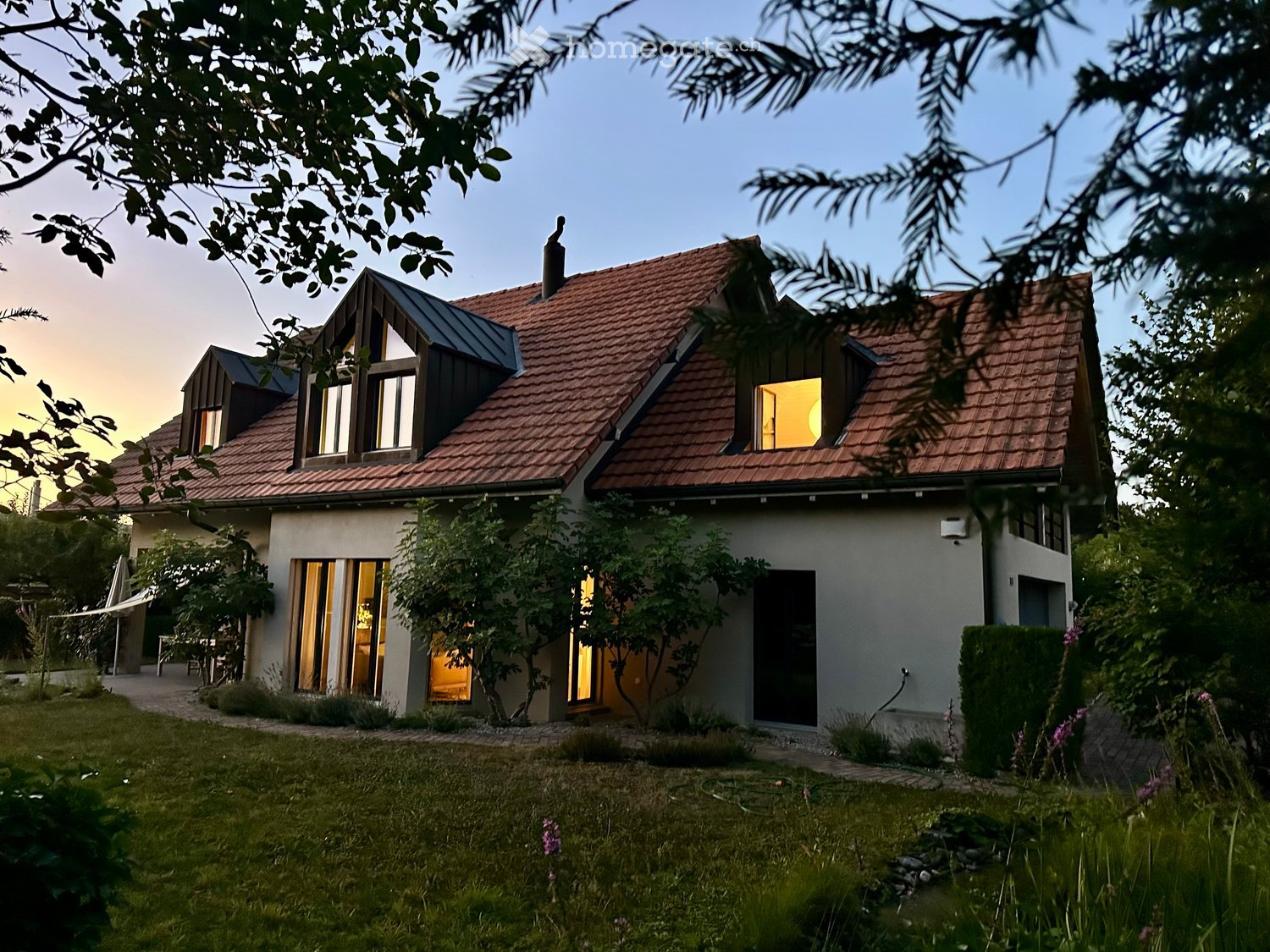 Two-story house with red-tiled roof, large windows, and modern design, surrounded by greenery and a well-maintained lawn.