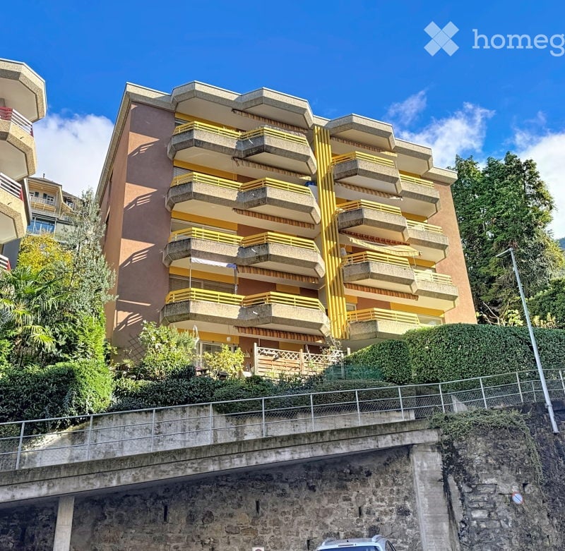 Multi-story residential building with balconies and yellow railings, situated on a raised ground floor with a stone retaining wall.