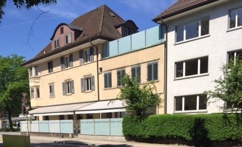 Multi-story residential building with a mix of modern and traditional architecture, featuring a combination of beige and white facades, large windows, and a blue balcony railing.