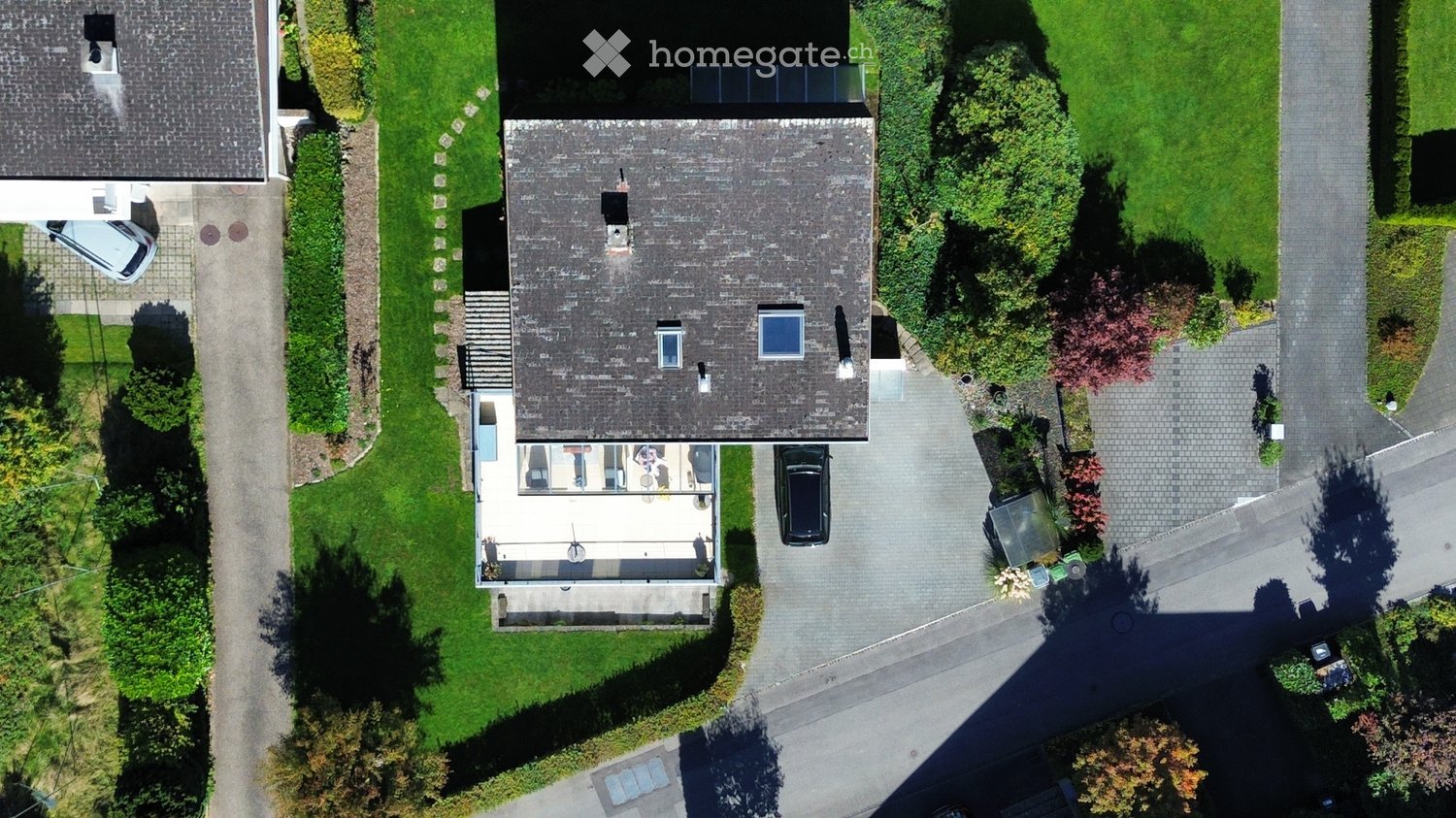 Aerial view of a two-story house with a grey roof, multiple windows, and a well-maintained garden. A driveway leads to a garage with a parked car, and there is a patio area with outdoor furniture.