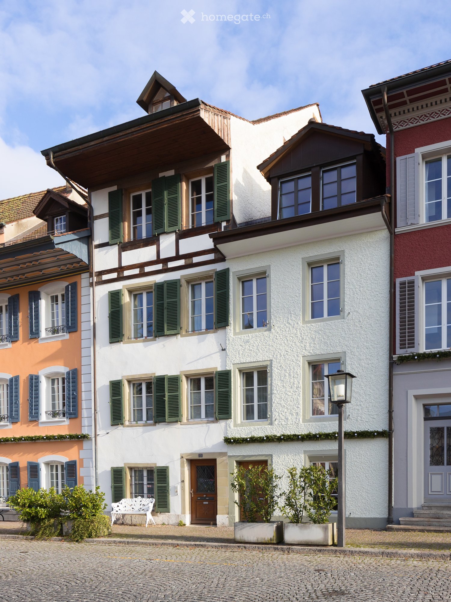 A two-story traditional house with white walls, green shutters, and a wooden balcony. The house features multiple windows, a central entrance, and is surrounded by small potted plants and a white bench.