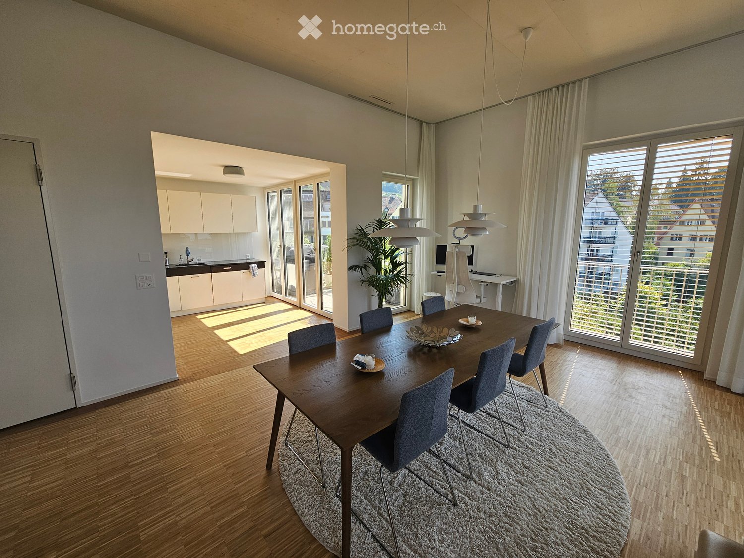 Open-plan dining and kitchen area with wooden flooring, large windows, and modern furniture including a dining table, chairs, pendant lights, and a small kitchen with cabinets and a sink.