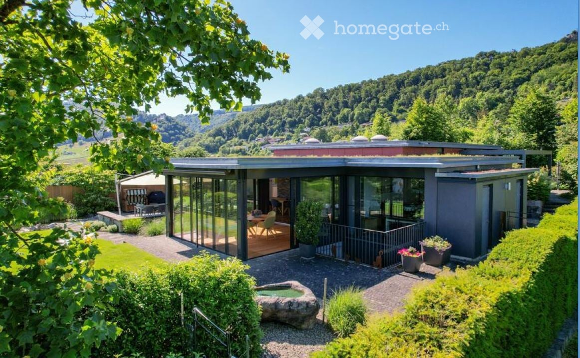 Modern house with glass walls, wooden deck, green roof, surrounded by greenery and mountains