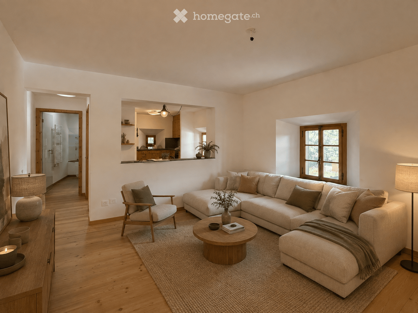 Modern living room with light-colored sofa, coffee table, armchair, floor lamp, and decorative items. Open-plan layout with visible kitchen area in the background.
