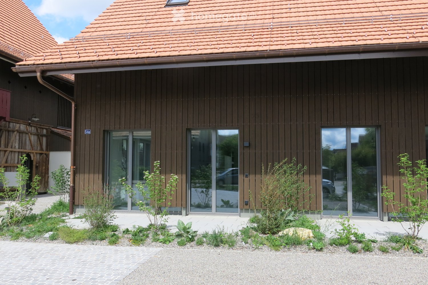 Modern two-story house with brown wooden facade, large glass windows, and a tiled roof. The front yard features small plants and shrubs arranged in a landscaped bed.