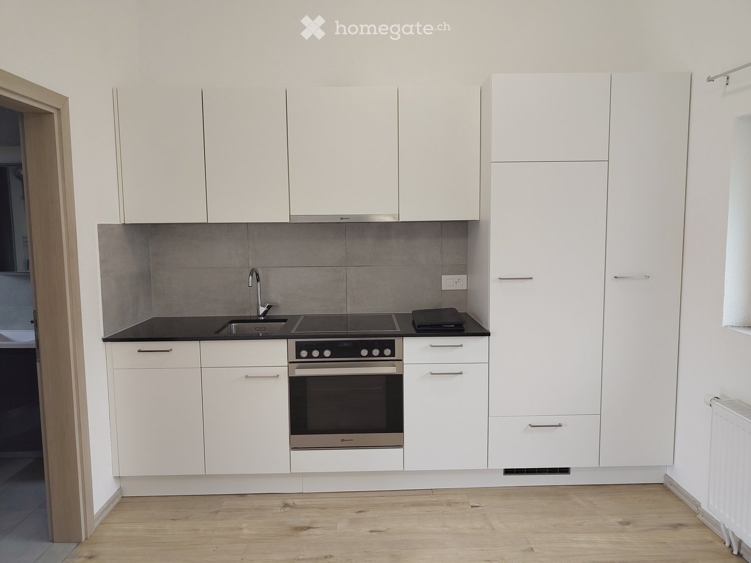 Modern kitchen with white cabinets, black countertop, stainless steel sink, stove, and oven. Wooden flooring and grey tiled backsplash.