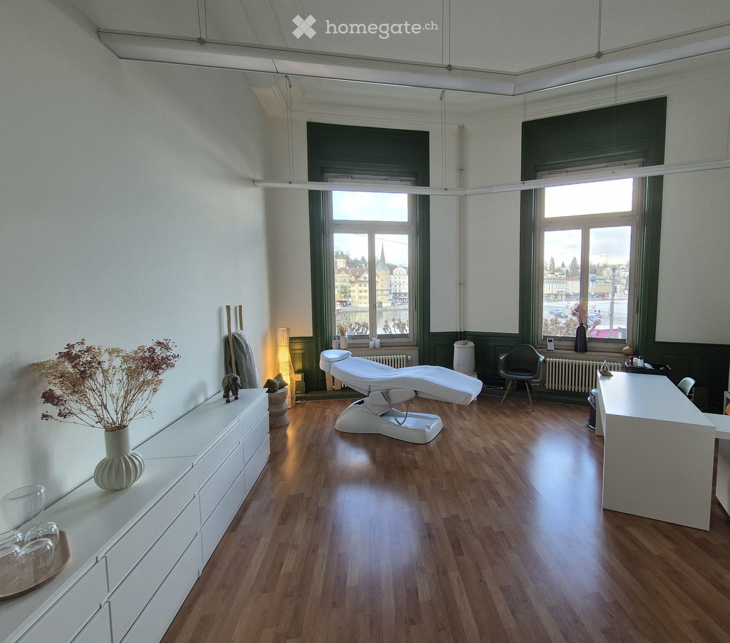 a room with wooden floor, 2 windows, a massage chair, a desk, a chair, a lamp, a vase with dried flowers, and a white cabinet with several drawers