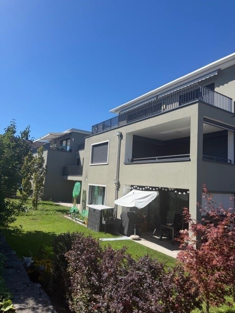Modern residential complex with balconies, outdoor seating area, umbrella, and well-maintained lawn.