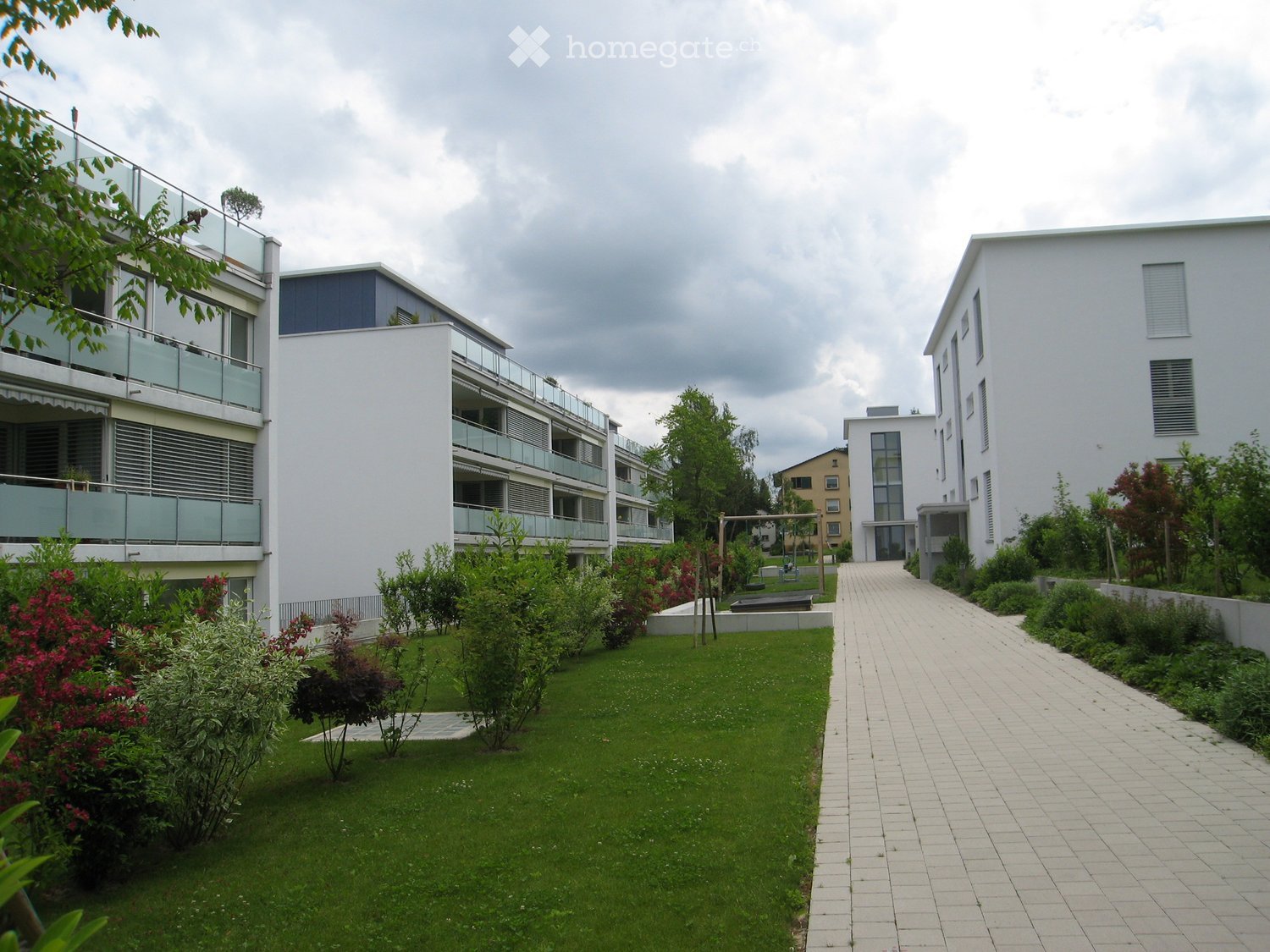 Modern residential complex with a well-maintained garden area, featuring a paved walkway, green lawn, shrubs, and trees. Balconies are visible on the buildings.