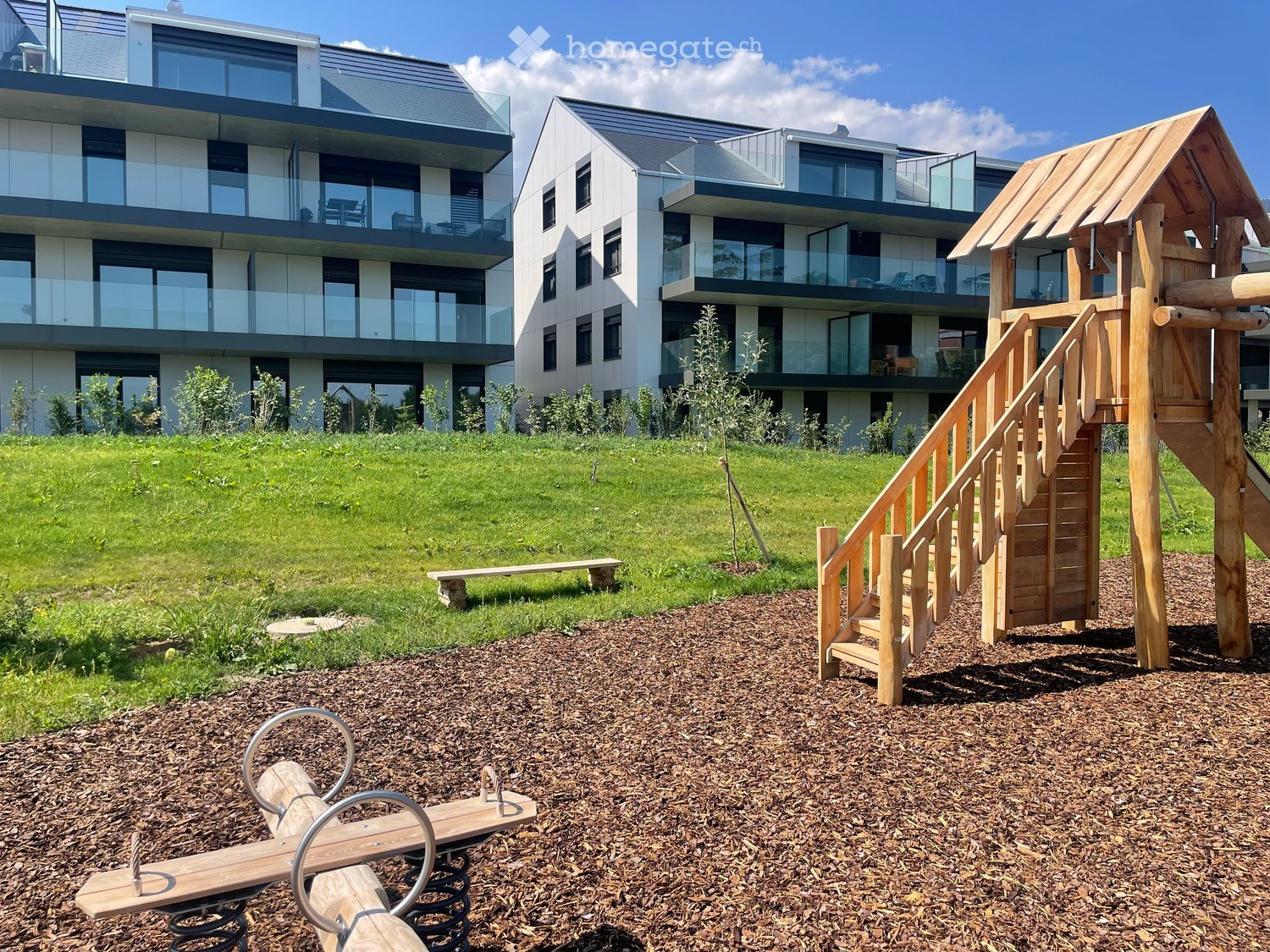 Modern residential complex with a playground area featuring a wooden playhouse, slide, and seesaw, surrounded by a grassy lawn and small trees.