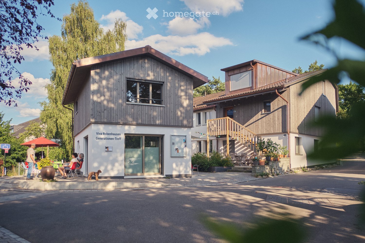 Two-story wooden building with a modern design, featuring large windows, a balcony, and a small outdoor seating area with tables and chairs.
