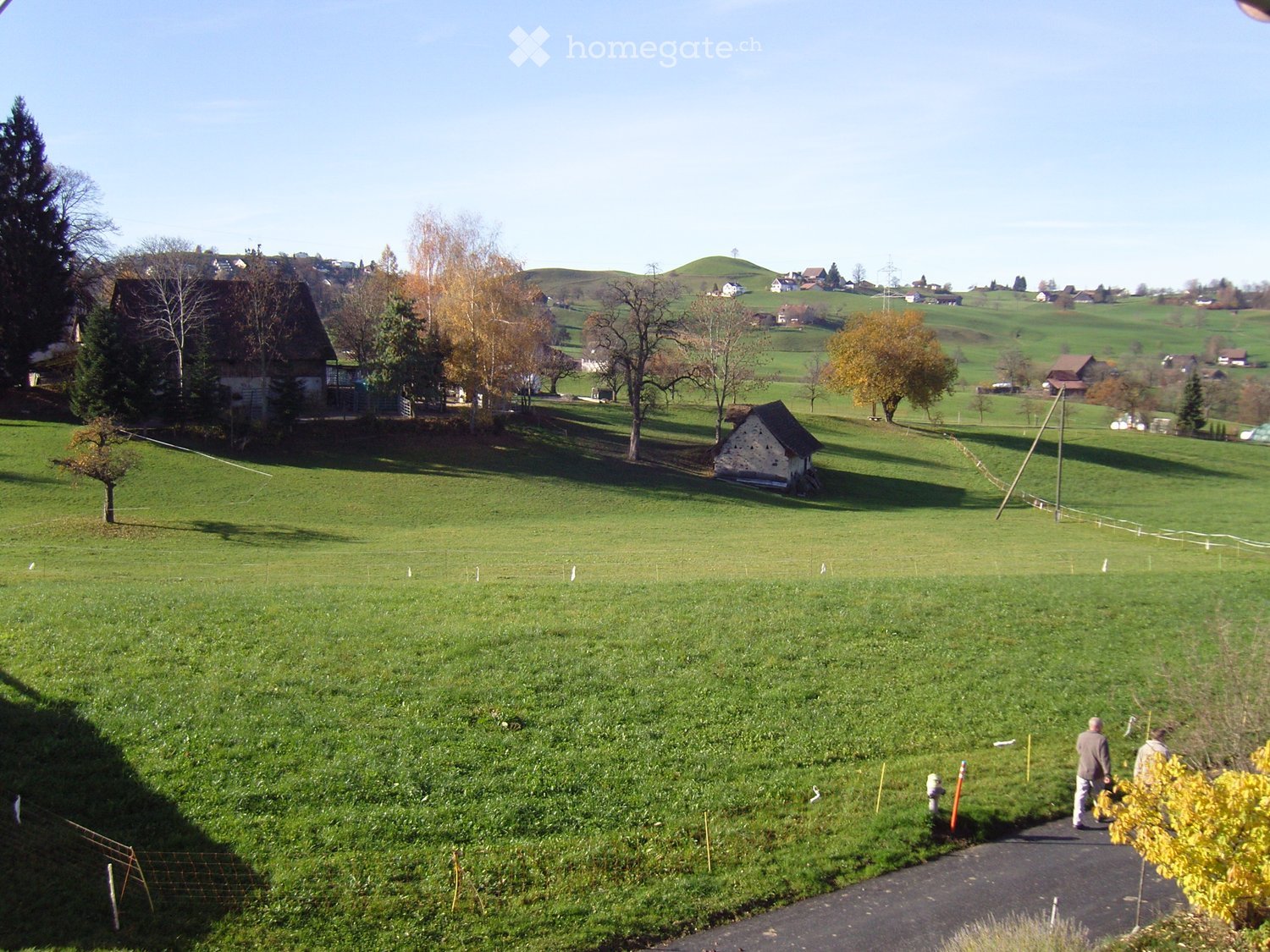 Rural landscape with green fields, scattered trees, and a few houses in the background. A paved path runs through the foreground with two people walking.