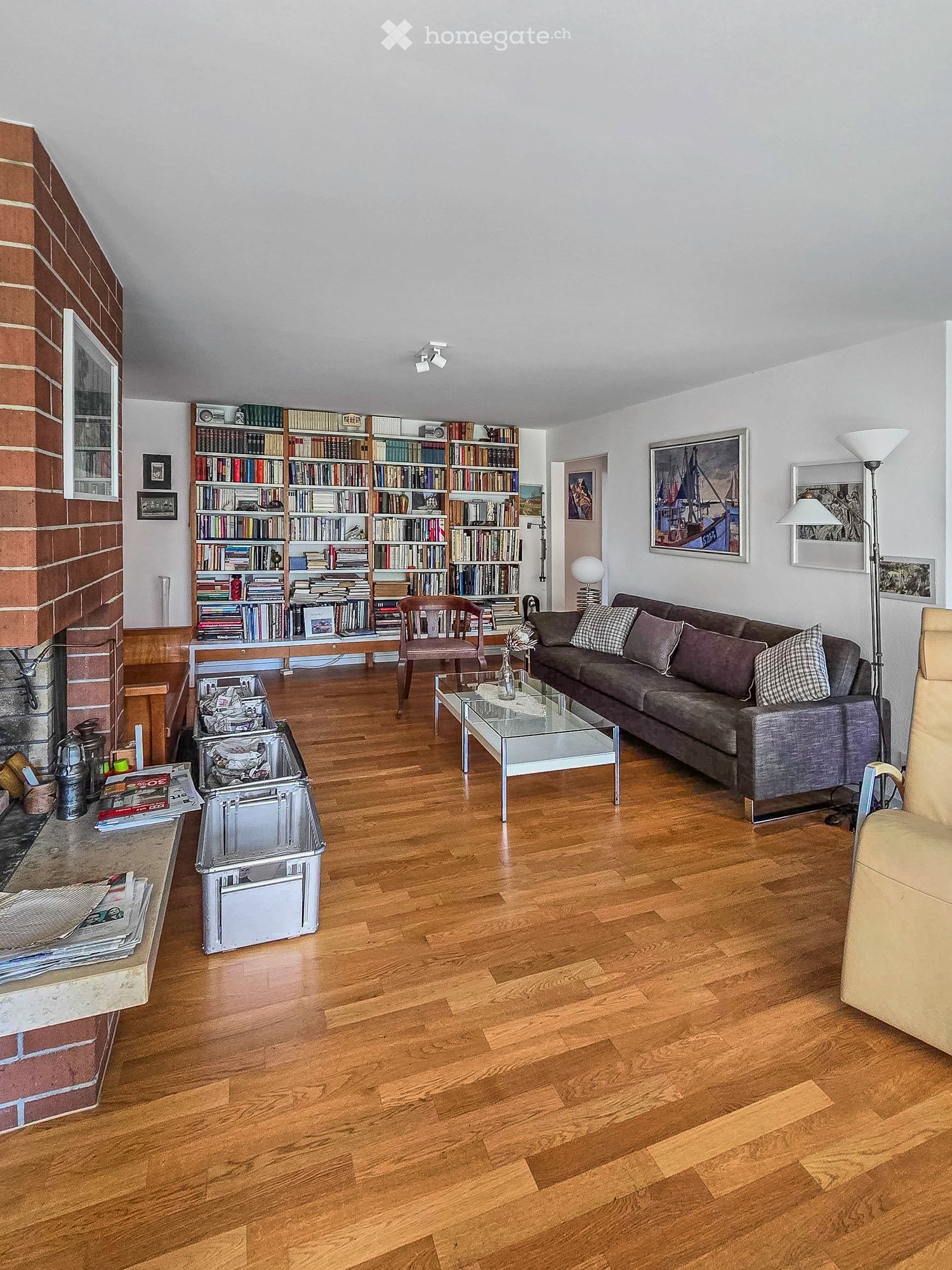 Living room with wooden flooring, brick fireplace, bookshelf, sofa, coffee table, and a chair.