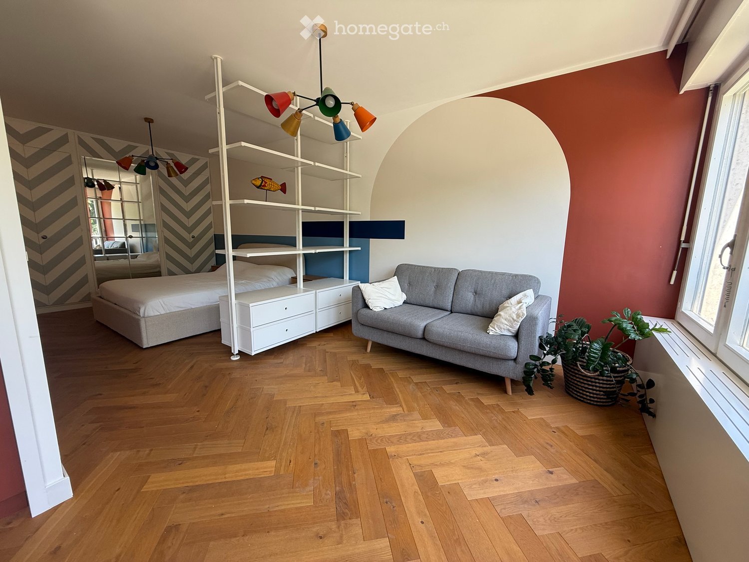 Modern furnished room with a grey sofa, bed, white shelving unit, and colorful pendant lights.