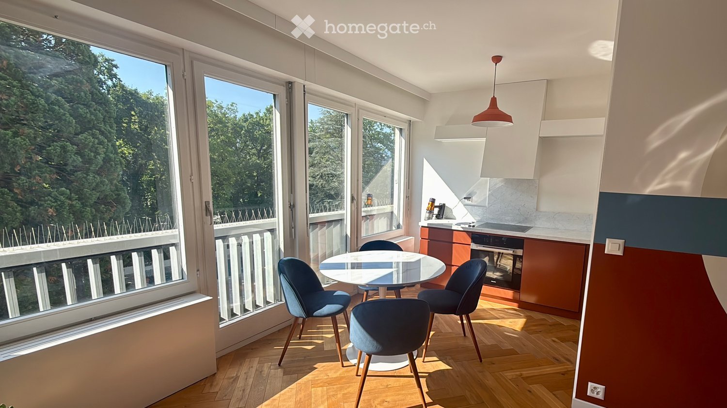 Modern, open-plan living and dining area with large windows overlooking a green view, a round glass dining table with four blue chairs, and a compact kitchen area with wooden cabinets and a built-in oven.