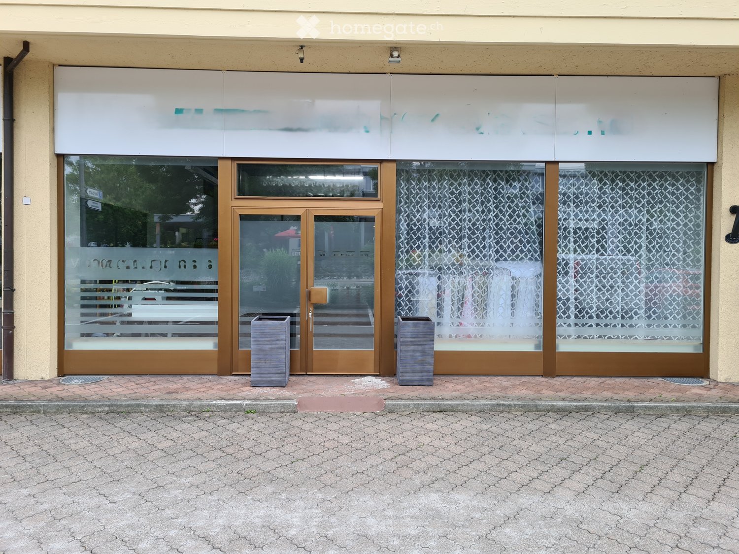 Facade of a commercial building with large glass windows, wooden-framed doors, and a modern signboard above. Two grey trash bins are placed in front of the entrance.