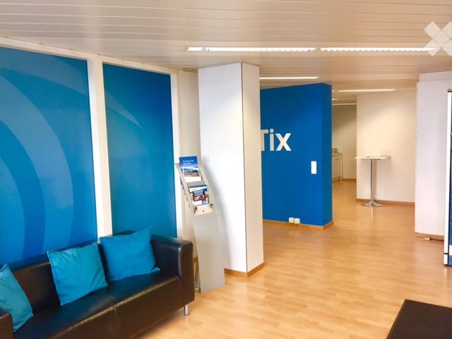 Modern reception area with a black leather sofa, a small table, and a screen stand. The walls are painted in blue with white trim, and the floor is made of light wood.