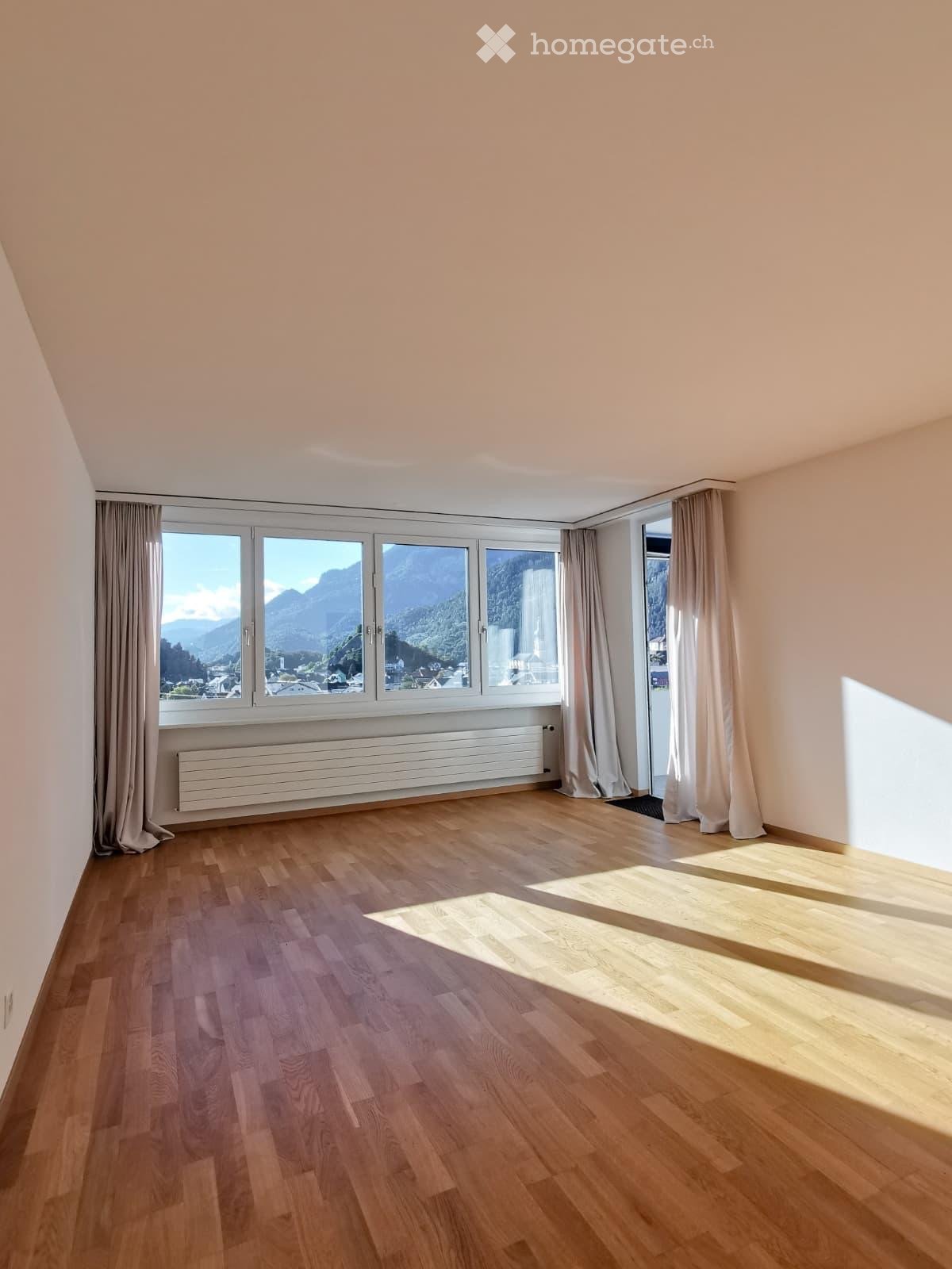 Empty room with wooden flooring, large windows with curtains, and a radiator below the windows.