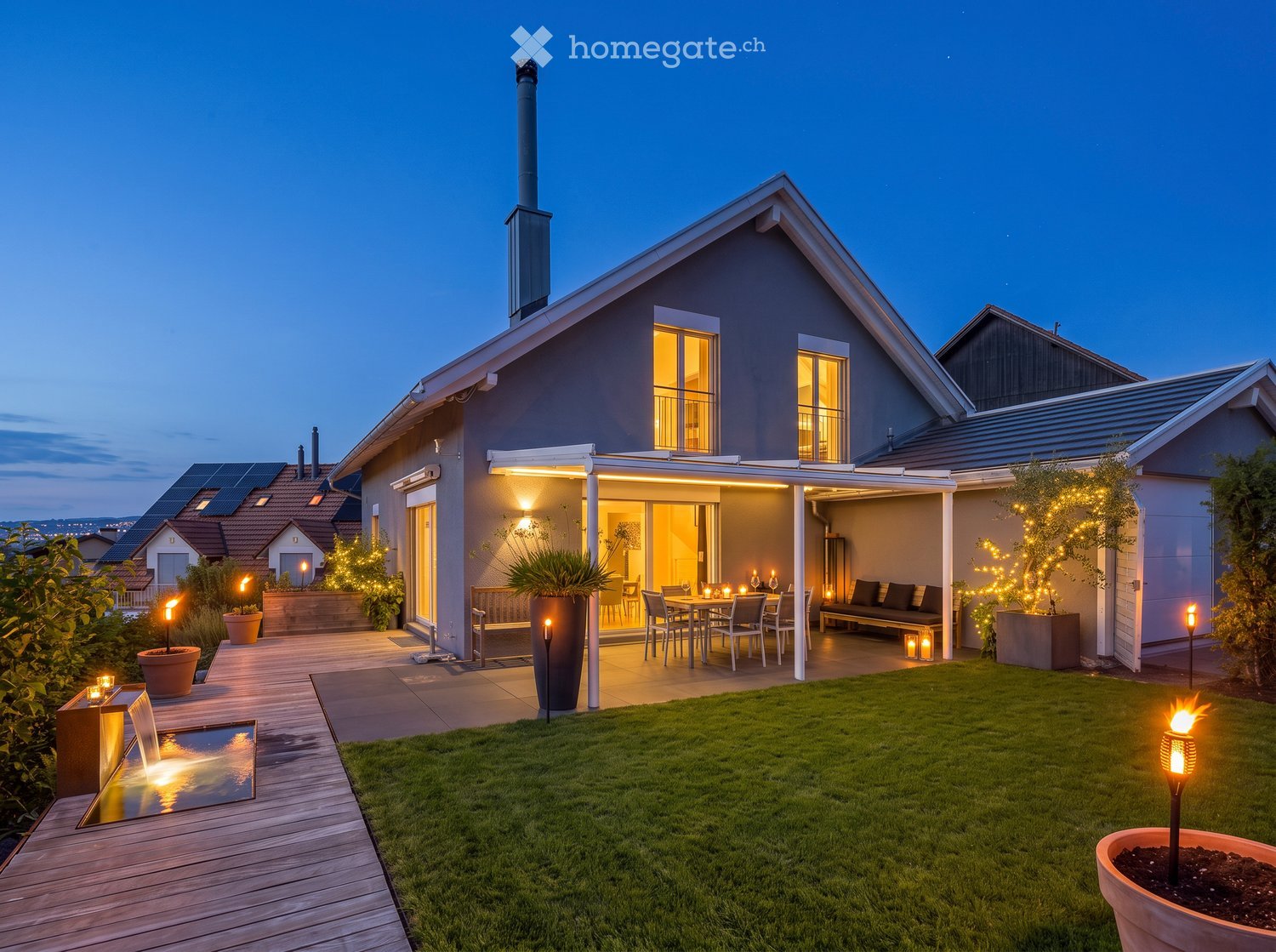 Well-maintained garden with a wooden deck, outdoor dining area, water feature, and ambient lighting.