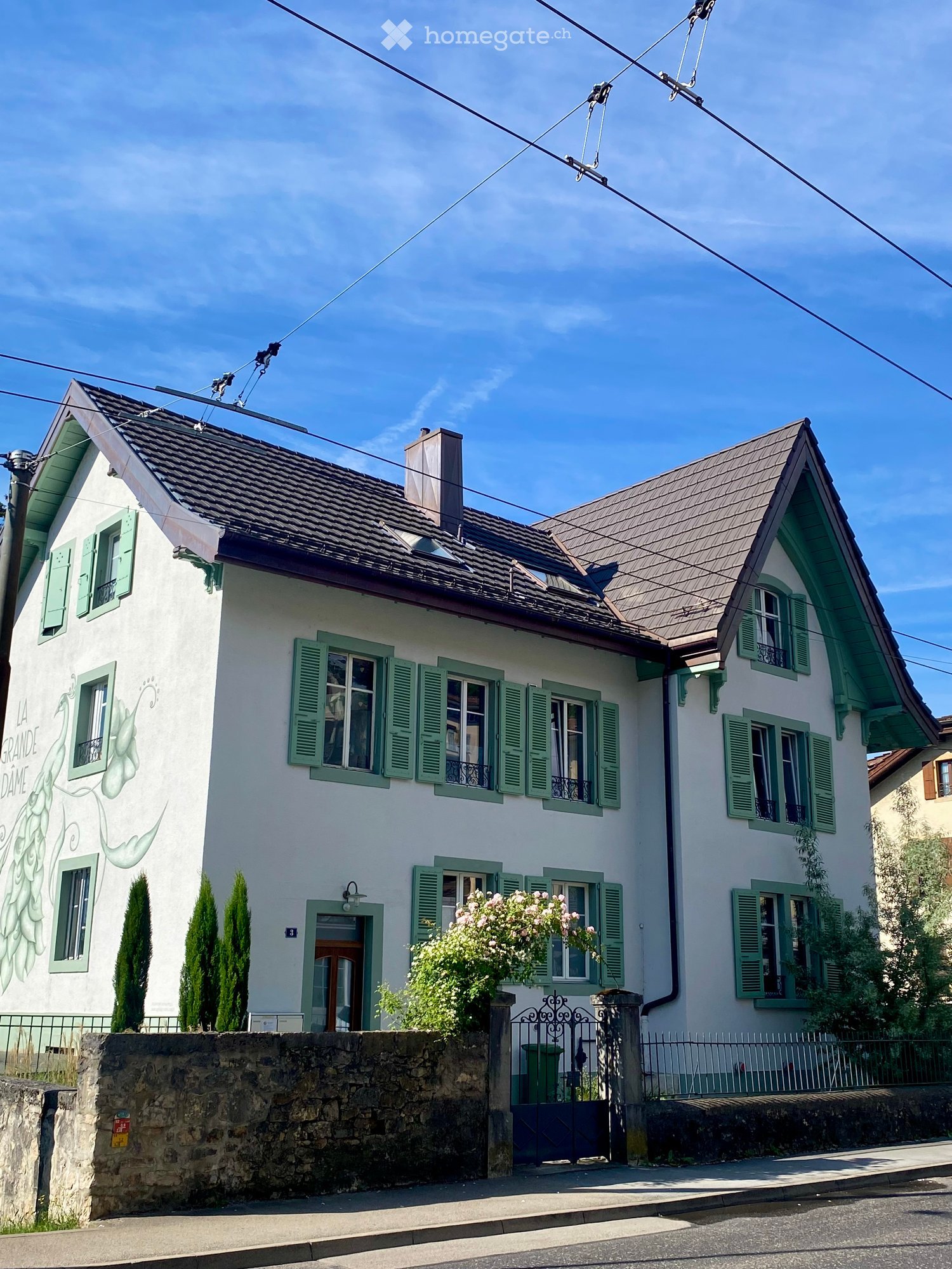 Two-story white house with green shutters, multiple windows, a chimney, and a decorative mural on the left side. The house has a stone wall in front and is surrounded by small trees and flowering bushes.