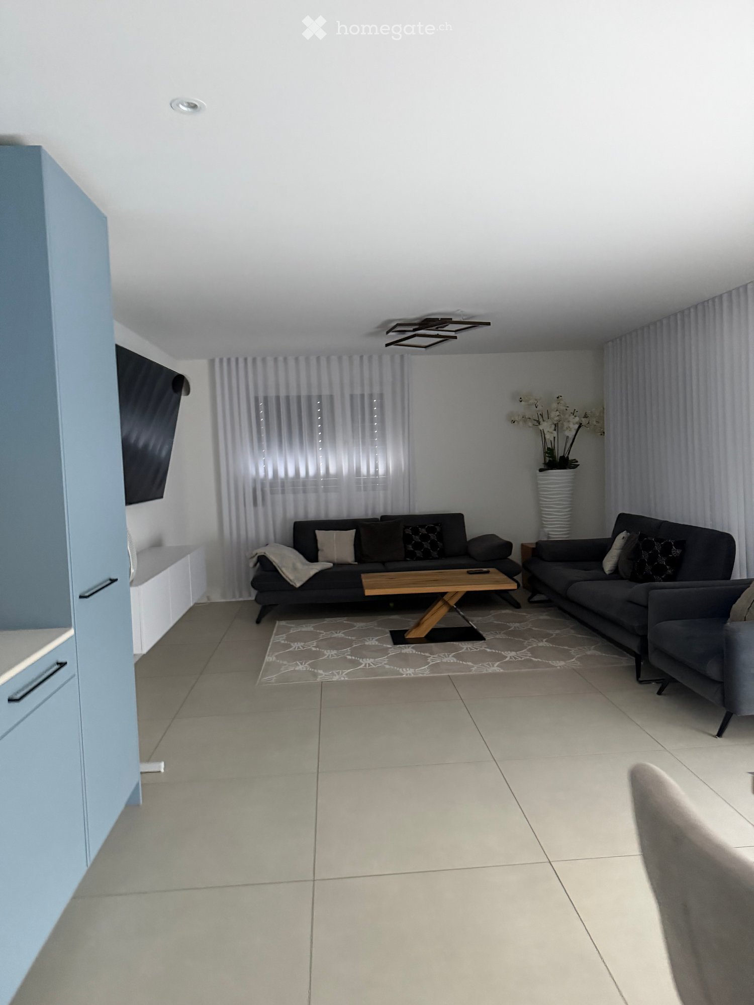 Modern living room with tiled floor, two sofas, coffee table, decorative vase, and large window with curtains.