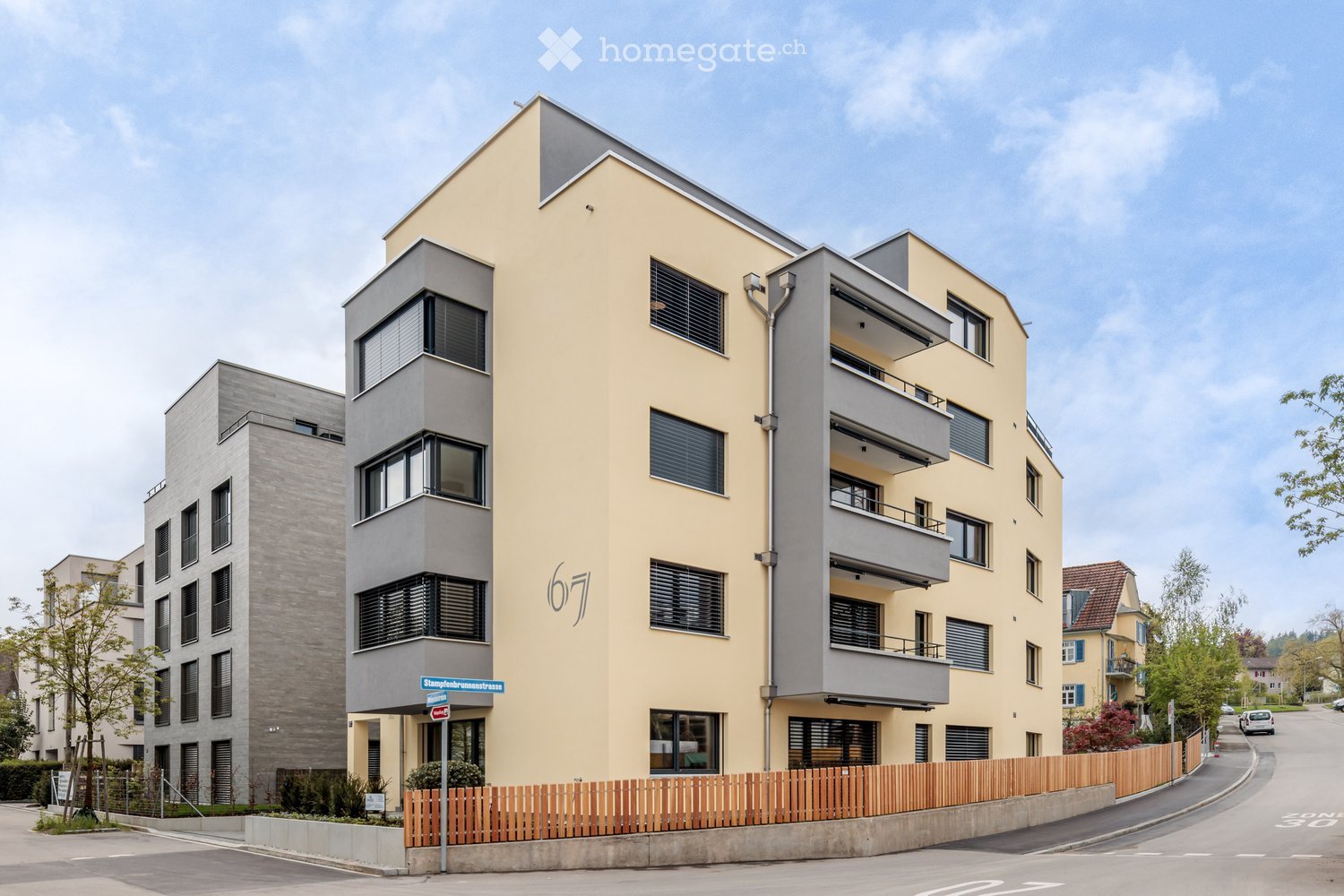 Modern multi-story residential building with a combination of light yellow and grey facades, large windows, and balconies. The building is situated on a street with a wooden fence and greenery in the foreground.