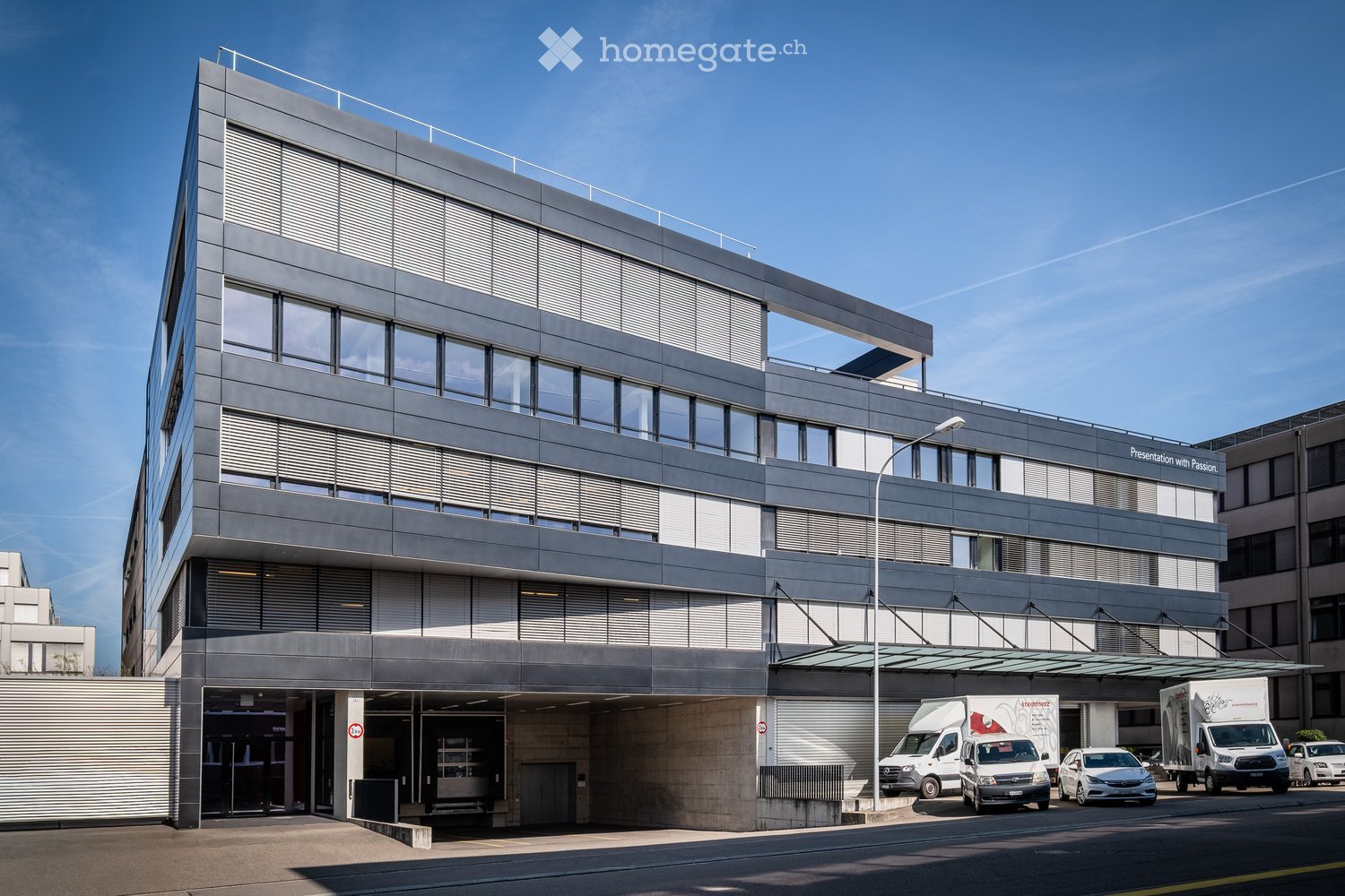Modern multi-story building with grey facade, large windows, and a covered entrance area. Several vehicles are parked in front.