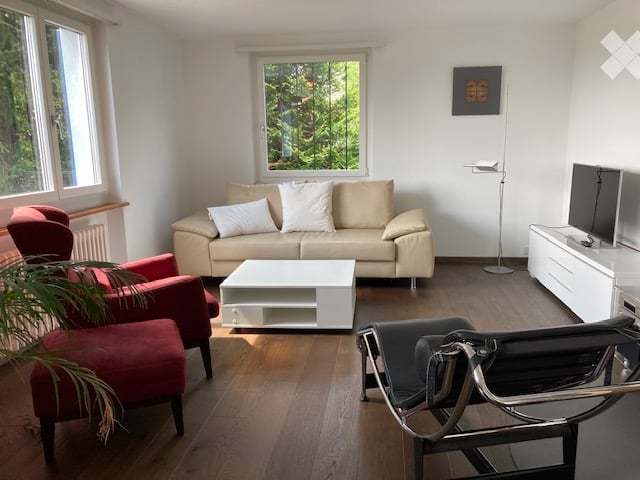 Bright living room with large windows offering a view of greenery, featuring a beige sofa, white coffee table, red armchairs, and a modern TV stand.