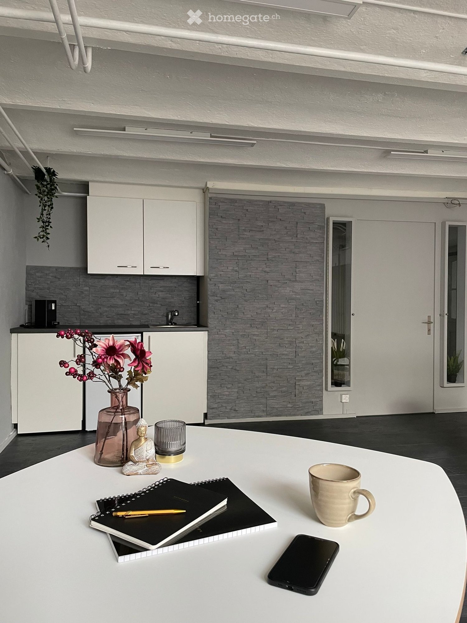 A modern, minimalist room with a white table holding a cup, cellphone, notebooks, pen, vase with flowers, and a small decorative figure. The room features white cabinets, a sink with a faucet, and a grey brick accent wall.