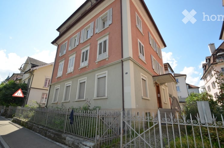A multi-story residential building with a red and cream facade, white shutters, and a metal fence in front.