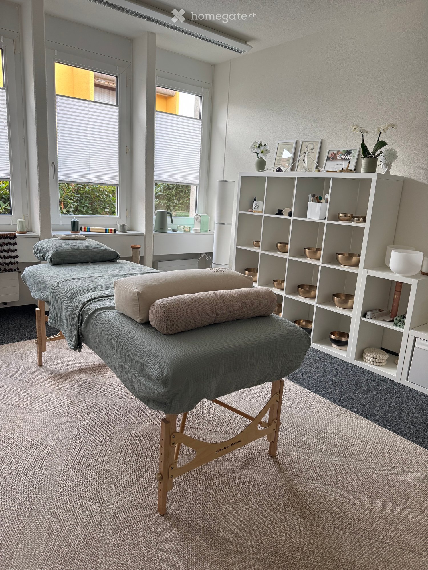 A serene, well-lit room with a massage table covered in a grey sheet and pillows, shelves with decorative items, vases, and framed pictures, and a large window with blinds.