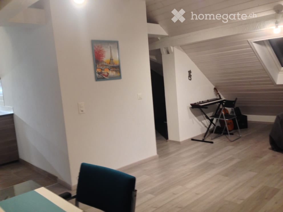A cozy attic room with wooden flooring, white walls, and a slanted ceiling. Features include a blue chair, a small table, a guitar, a keyboard on a stand, and a picture frame on the wall.
