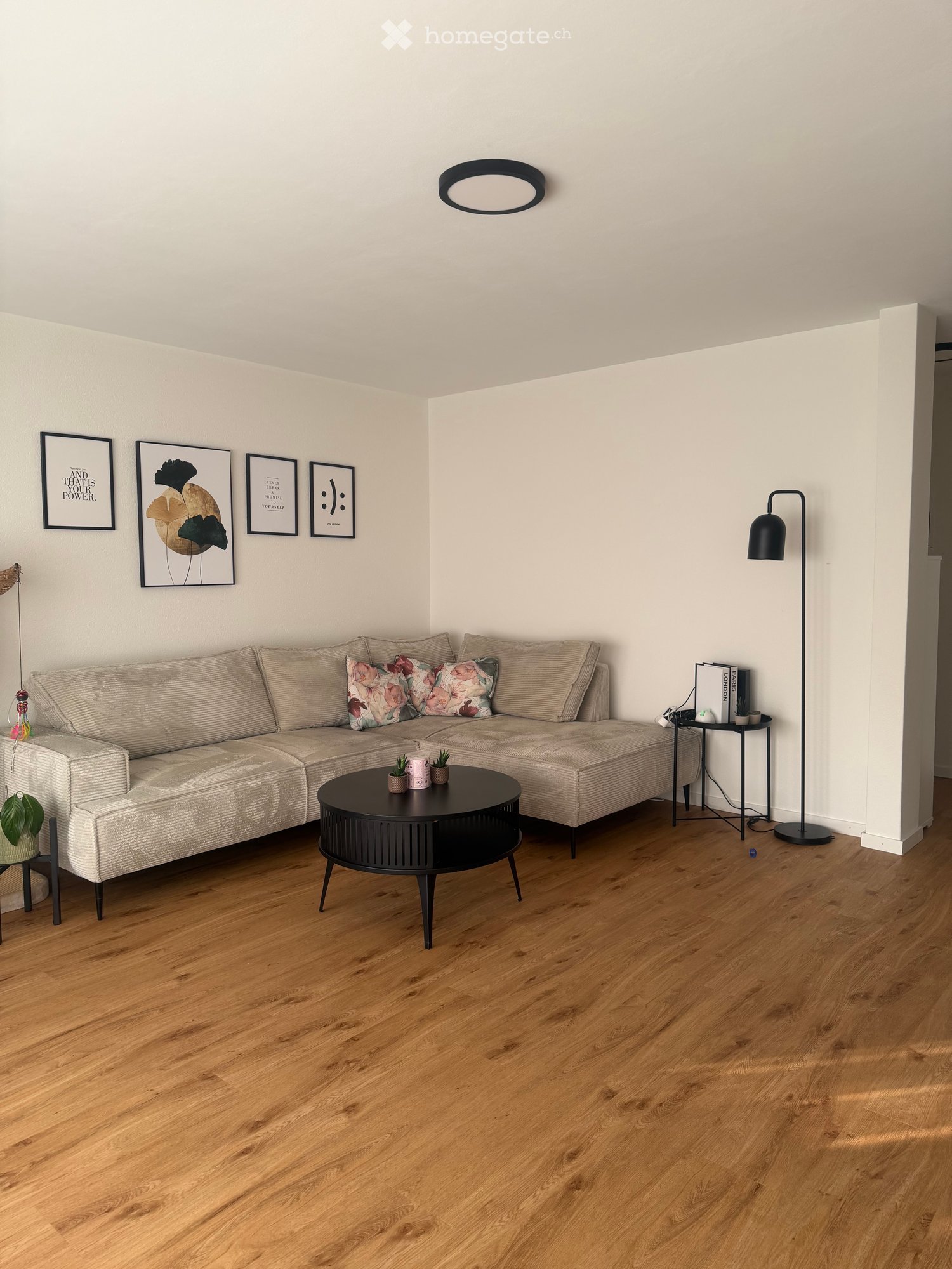 Modern living room with light-colored sectional sofa, coffee table, floor lamp, side table, potted plants, and picture frames on the wall.