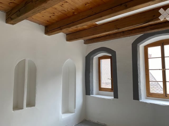 Empty room with white walls, wooden ceiling beams, and arched windows, including one with wooden shutters.