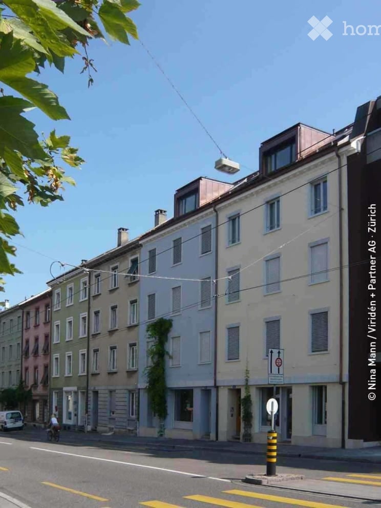 Multi-story residential building with multiple windows and balconies, located on a street with a clear blue sky and some greenery visible.