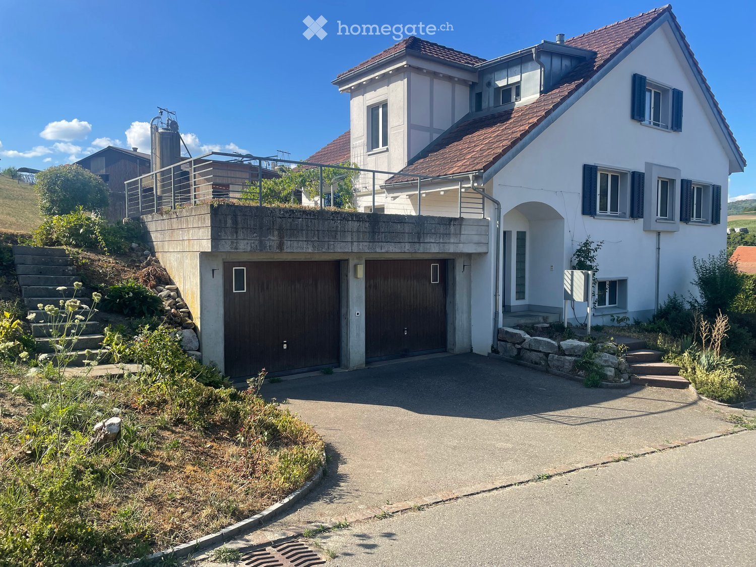 Two-story house with a raised ground floor, garage, and balcony on the upper level. The house has white walls, dark shutters, and a red-tiled roof.