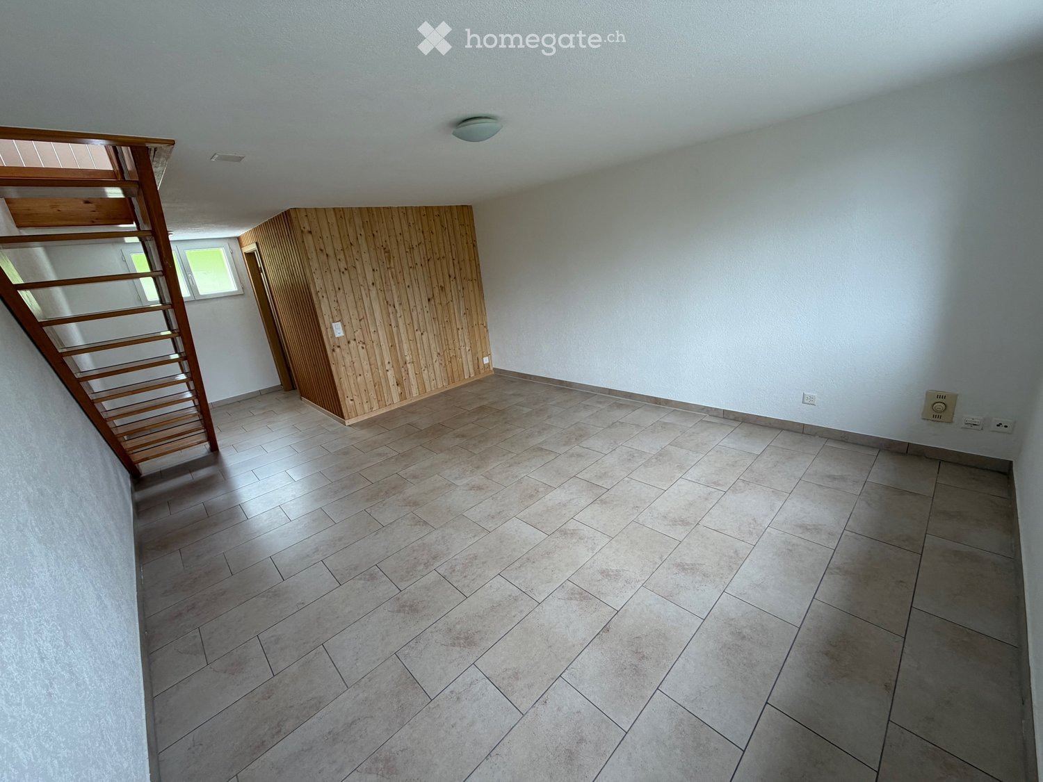 Empty room with tiled floor, white walls, and a wooden staircase leading upwards.
