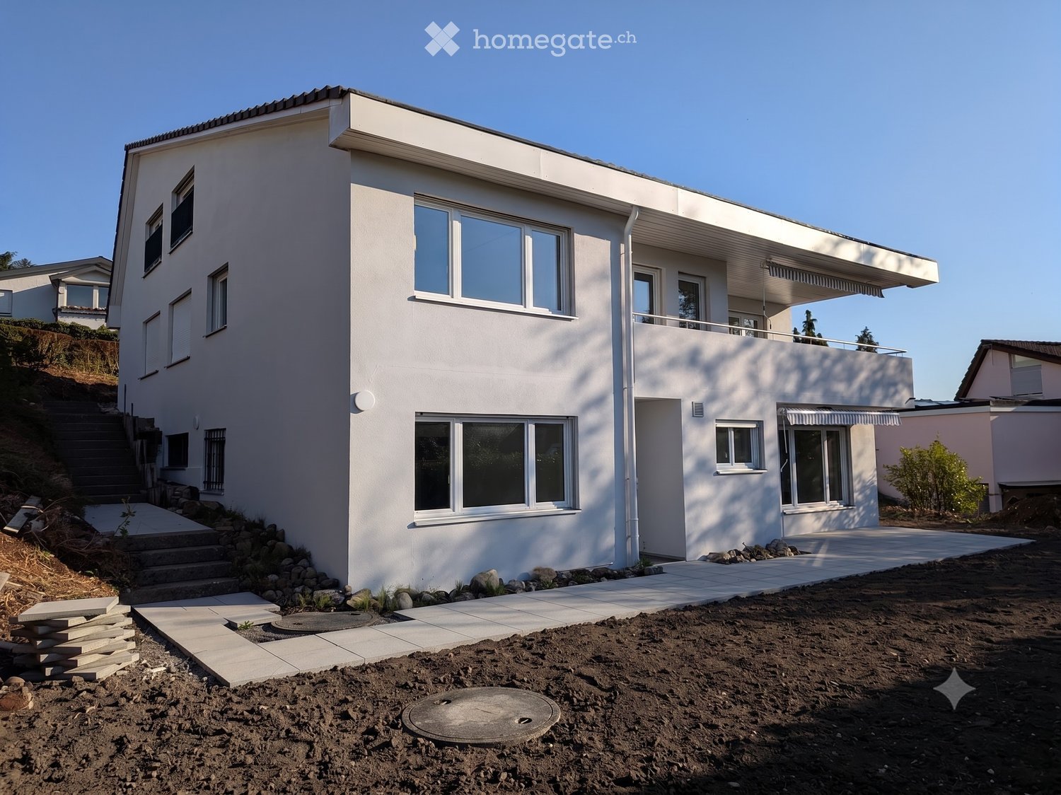 Modern two-story house with white exterior, large windows, and a raised ground floor entrance. Features include a balcony, steps leading up to the house, and a partially constructed patio area.