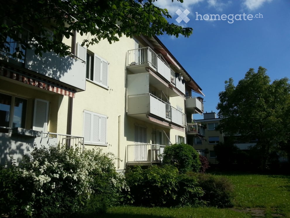 Multi-story residential building with balconies, white shutters, and greenery in front.
