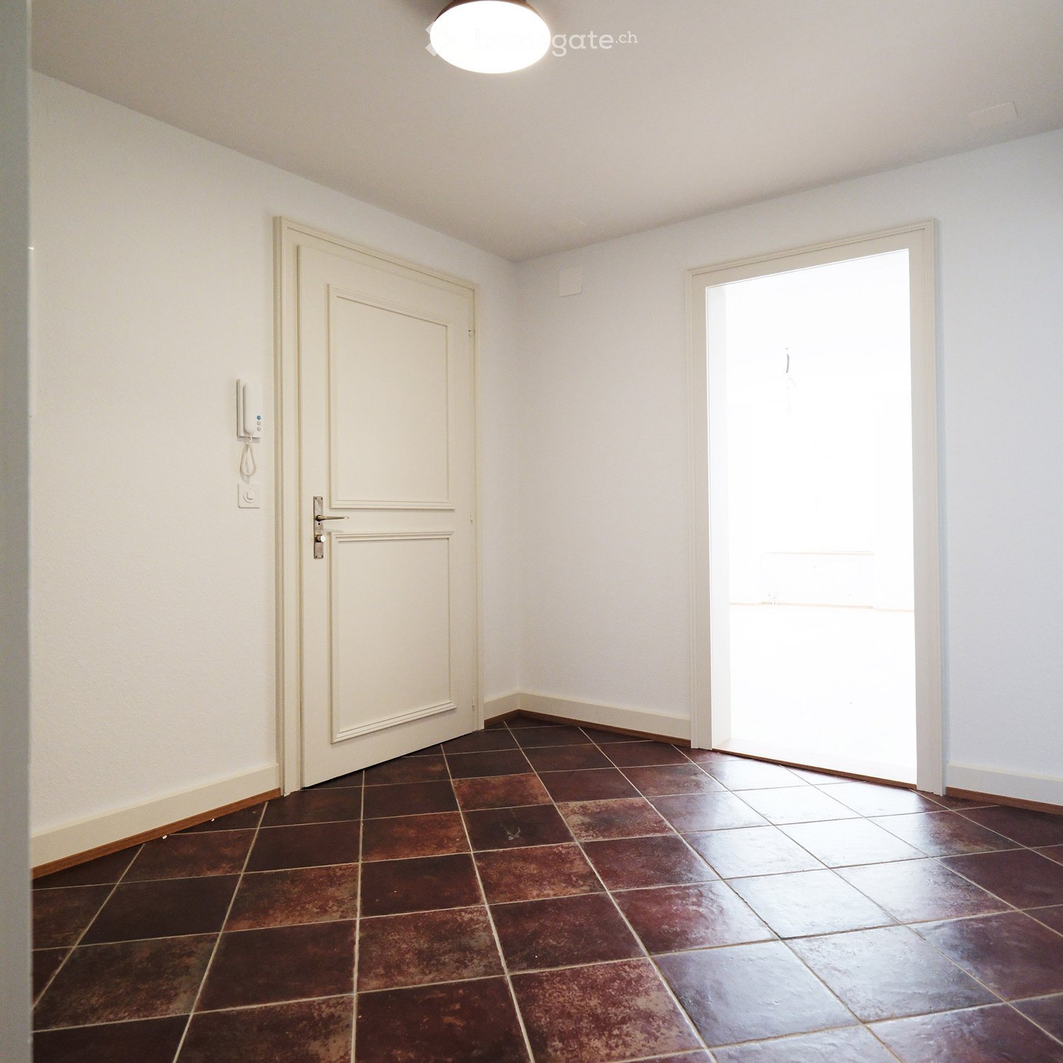 Empty hallway with brown tiled floor, white walls, and a closed door on one side and an open door on the other.
