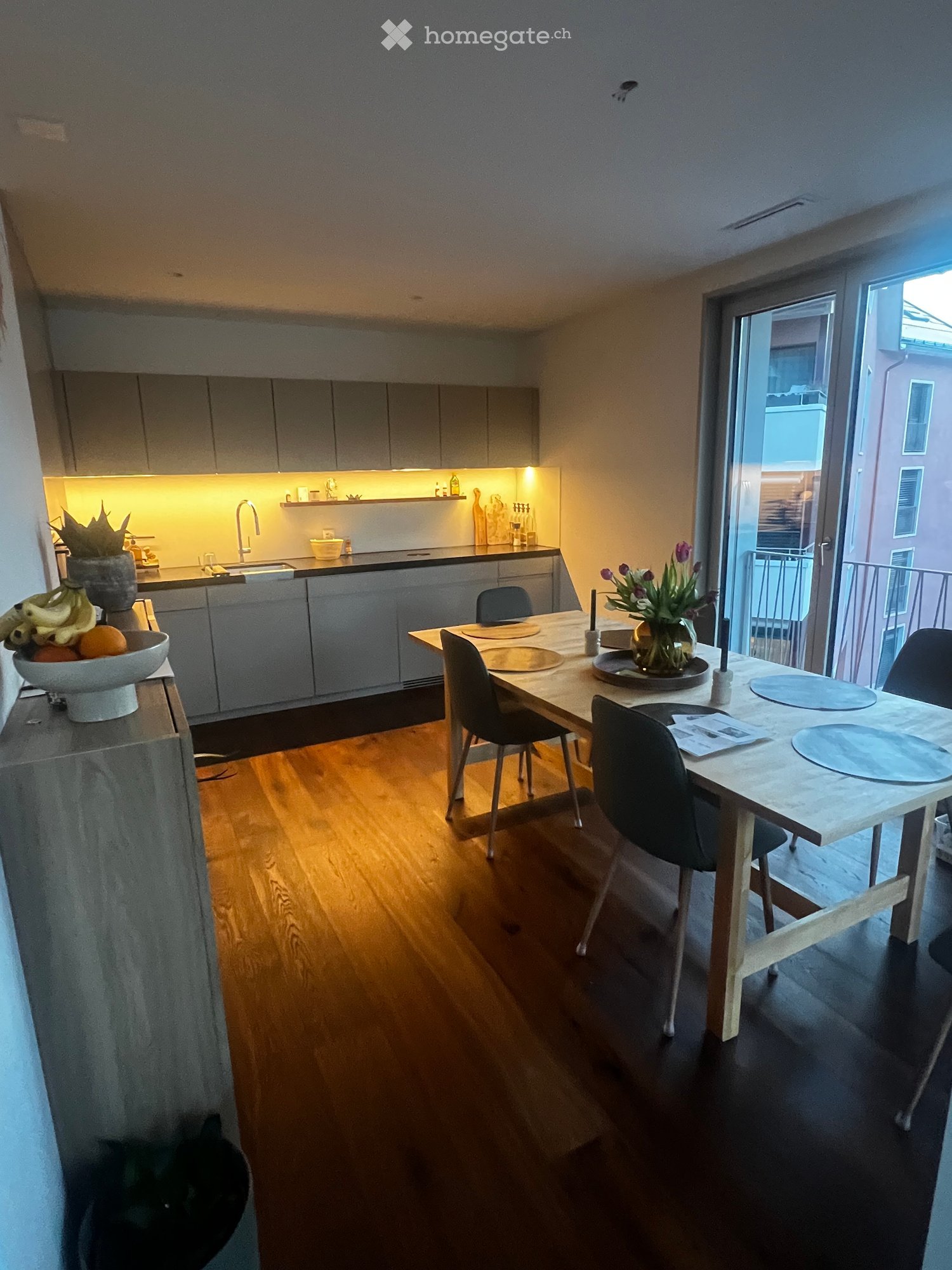 Modern kitchen and dining area with wooden flooring, grey cabinets, a dining table with chairs, and a vase of flowers on the table.