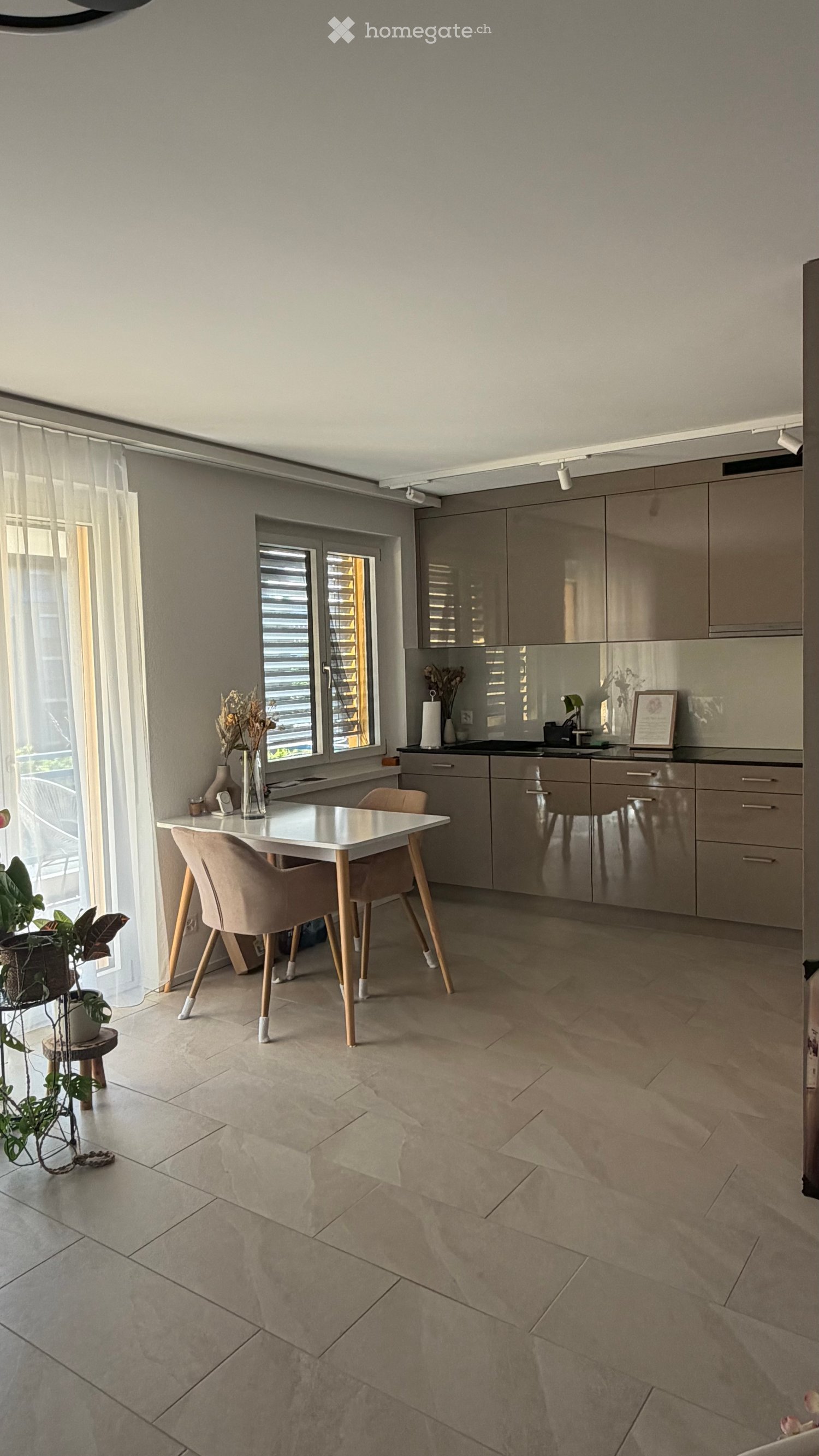 Modern kitchen with light grey cabinets, a white dining table with four chairs, a sink, and decorative items including a framed picture and vases. The space features large windows with blinds and a tiled floor.