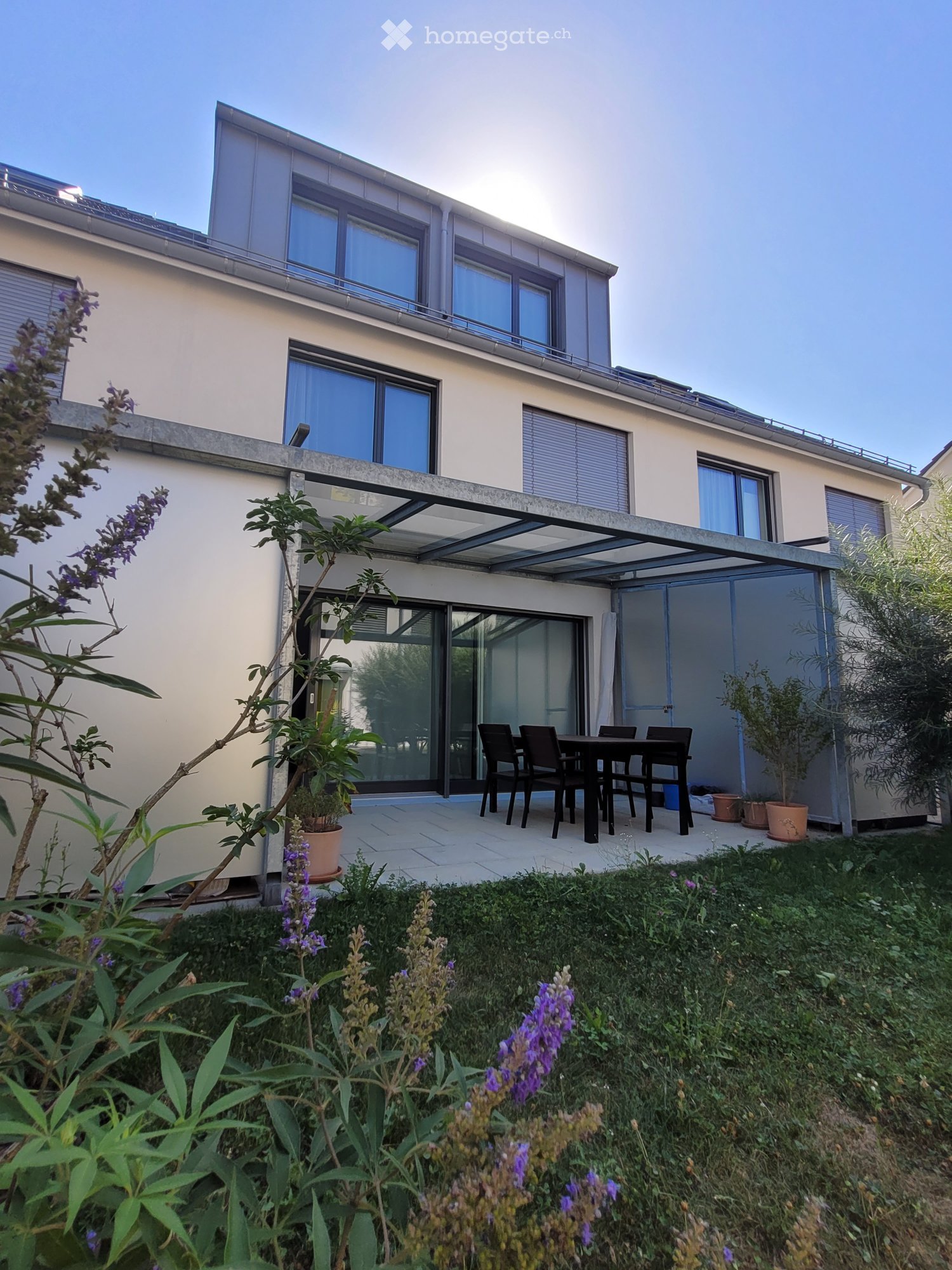 Modern two-story house with large windows and a covered patio area. The patio features a black dining table and chairs, surrounded by potted plants and lush greenery.