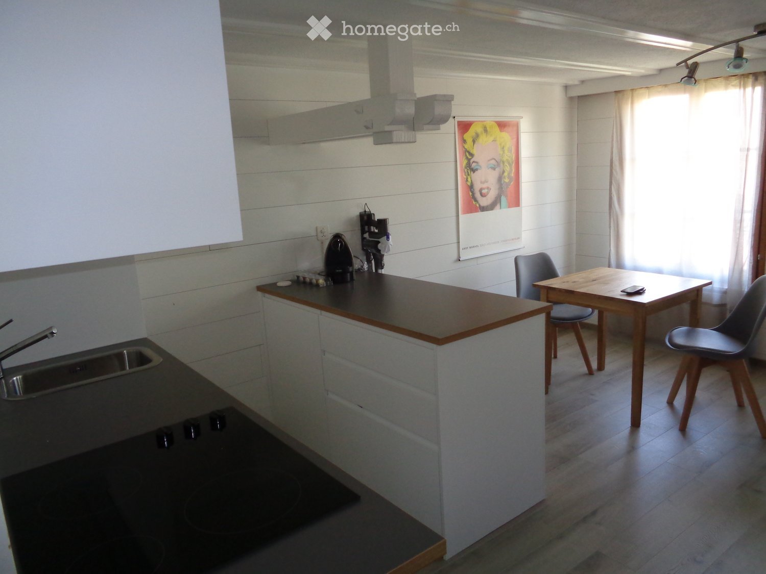 Modern kitchen with a sink, faucet, coffee maker, and a small dining area with a table and chairs. The room features white shiplap walls, wooden flooring, and a large window with white curtains.